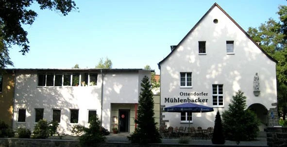 Zwei Gebäude mit der Aufschrift Ottendorfer Mühlenbäcker, vorne ein Cafébereich mit Tischen und Sonnenschirm.