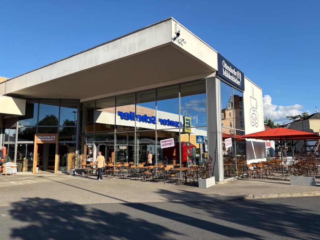 Moderne Bäckereifassade mit großen Glasfenstern und Außenterrasse; Personen gehen vorbei, blauer Himmel im Hintergrund.