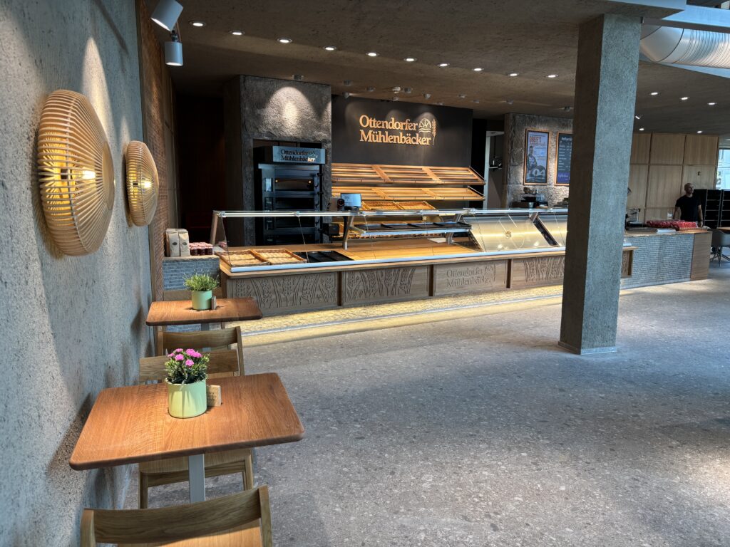 Bäckerei-Innenraum mit leerer Verkaufstheke, drei Holztischen mit Blumen und an der Wand montierten Lampen.
