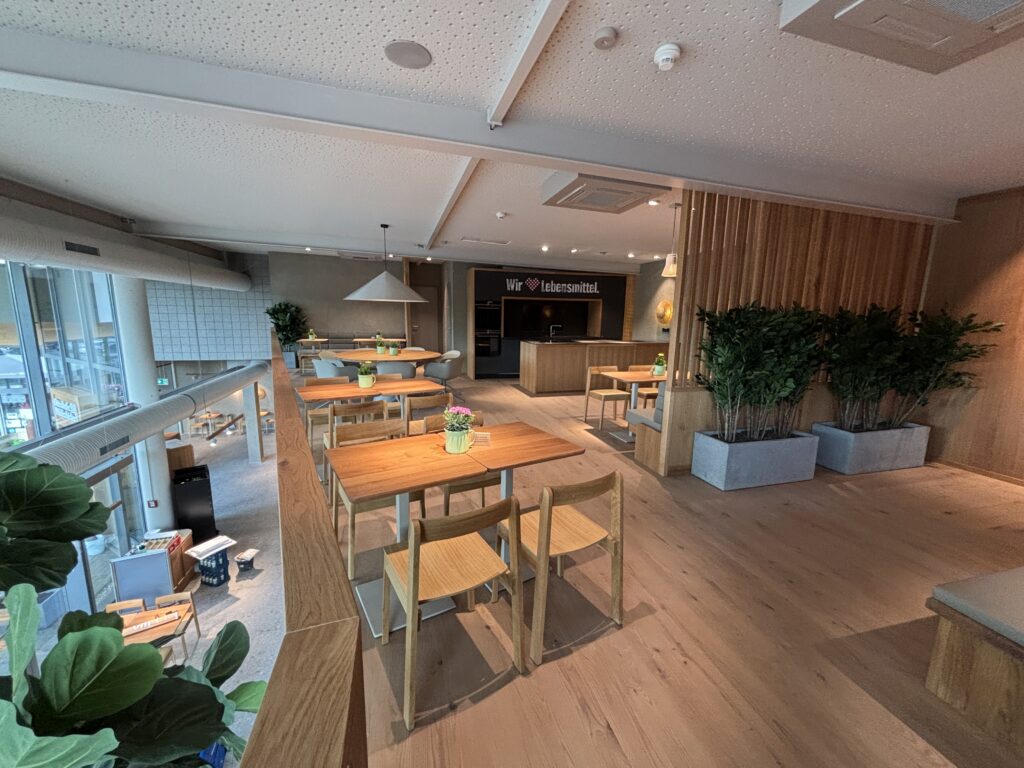 Moderner Cafébereich mit Holztischen, -stühlen, Pflanzen und großen Fenstern. Im Hintergrund ein Schild: Wir ♥ Lebensmittel.