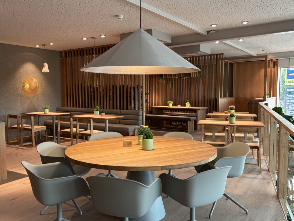 Moderner Café-Innenraum mit Holztischen, grünen Stühlen und Pflanzen auf den Tischen, große Leuchte in der Mitte.